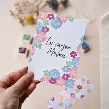 Mano sosteniendo una postal floral "La mejor mama", diseñada con colores suaves y detalles botánicos